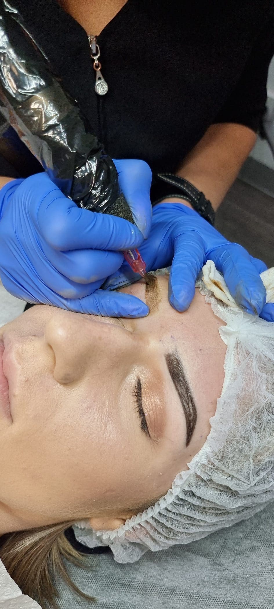 A technician wearing blue gloves uses a microblading pen to tattoo a woman's eyebrow during a cosmetic procedure.