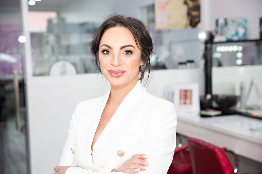 A smiling woman with dark hair and a white blazer posing with crossed arms in a modern beauty clinic.