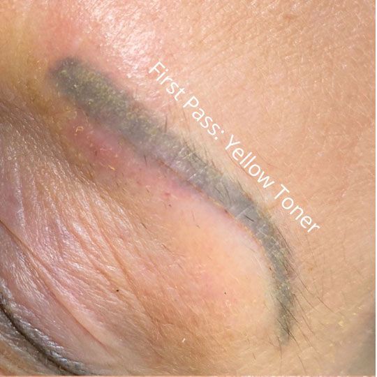 Close-up of an eyebrow with a bluish-grey tattoo being treated with a yellow toner, with the text 'First Pass: Yellow Toner' visible.