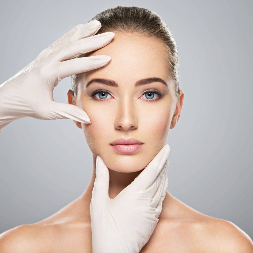 A woman with blue eyes and light skin is having her face examined by gloved hands, likely for a cosmetic consultation.