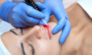 A close-up shows a technician in blue gloves applying permanent red lip makeup to a woman with a tattooing machine.