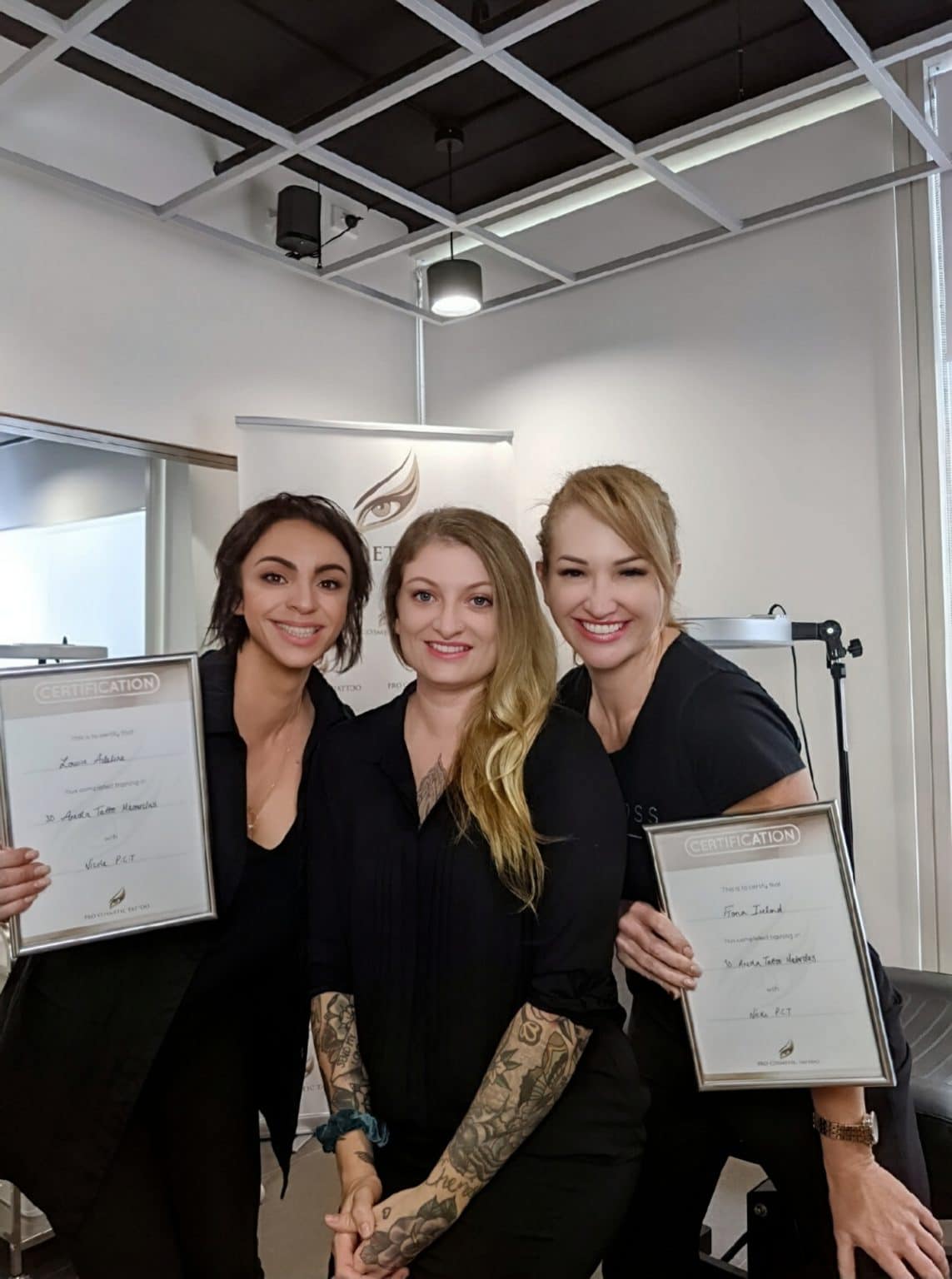 Three smiling women, two holding framed certificates for a '3D Areola Tattoo Masterclass', standing indoors.