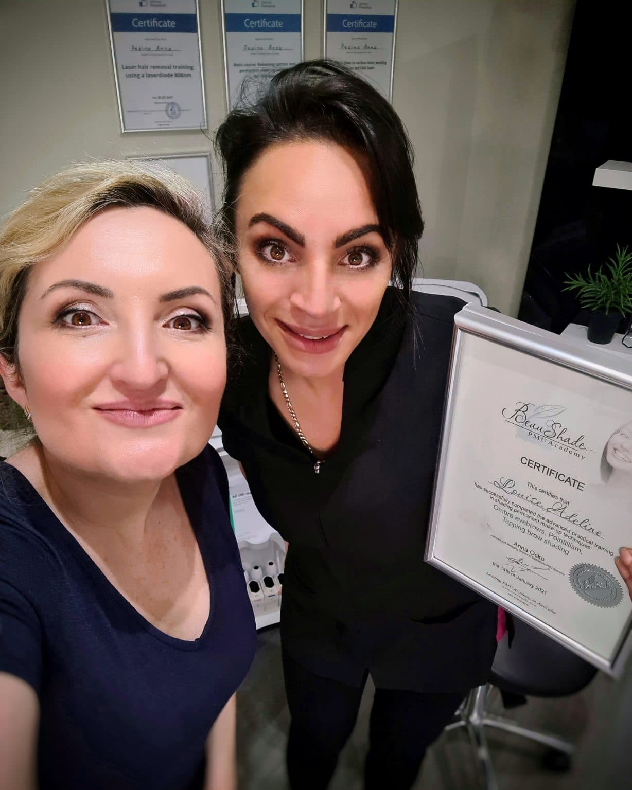 Two smiling women, one with blonde hair and the other with dark hair holding a "Beau Shade PMU Academy" certificate, stand in a room with other certificates on the wall.